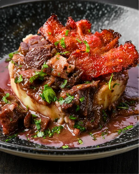 Langzaam gestoofd vlees met een knapperige rode korst ligt bovenop romige aardappelpuree in een donker gevlekt kommetje, omringd door een rijke wijnsaus en verspreide verse kruiden.