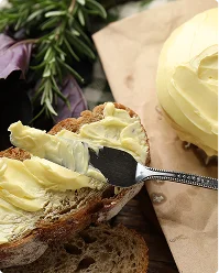 Een zilveren mes smeert een royale laag romige boter op een dikke snee donker volkorenbrood. Een rond klontje boter en een takje verse rozemarijn liggen ernaast op bruin bakpapier.