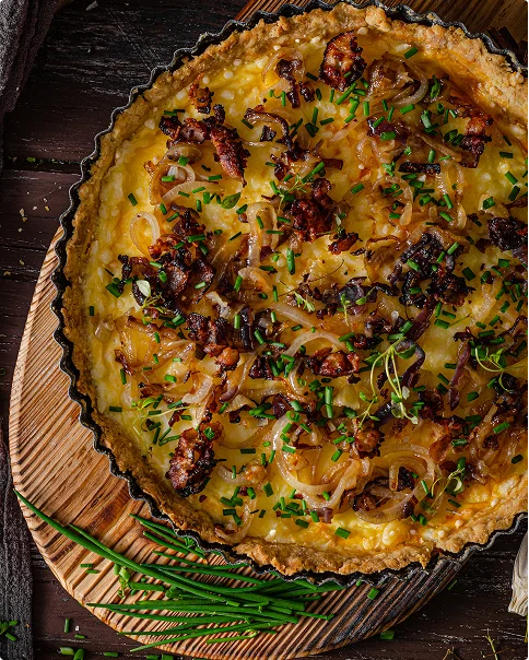 Een gouden quiche in een geribbelde taartvorm is bedekt met gekarameliseerde uien, knapperige stukjes en vers gesneden bieslook, rustend op een ronde houten plank met een bosje bieslook erbij.
