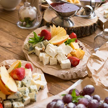 Een houten tafel draagt verschillende kaas- en fruitplateaus met diverse kazen, aardbeien, appelpartjes, druiven en verse kruiden, naast een pot jam en glaswerk.