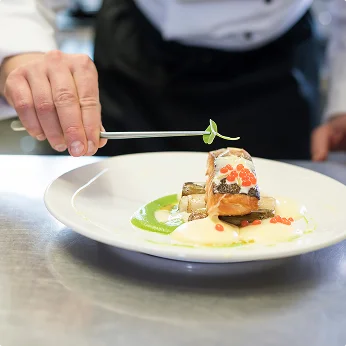 De hand van een chef gebruikt een pincet om een klein kruidengarnituur bovenop een fijnproeversgerecht van gelaagde vis met rode kaviaar en groene saus te plaatsen.