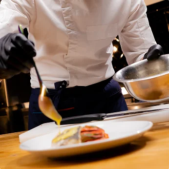 Een chef-kok in een wit jasje en zwarte handschoenen schept gouden saus uit een pan op een bord in een professionele keuken.