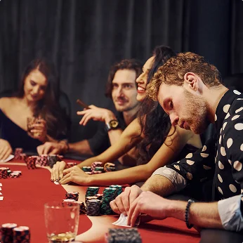Een groep jonge volwassenen zit rond een rood vilten pokertafel met stapels fiches, drankjes in de hand, lachend en kaartspelend.