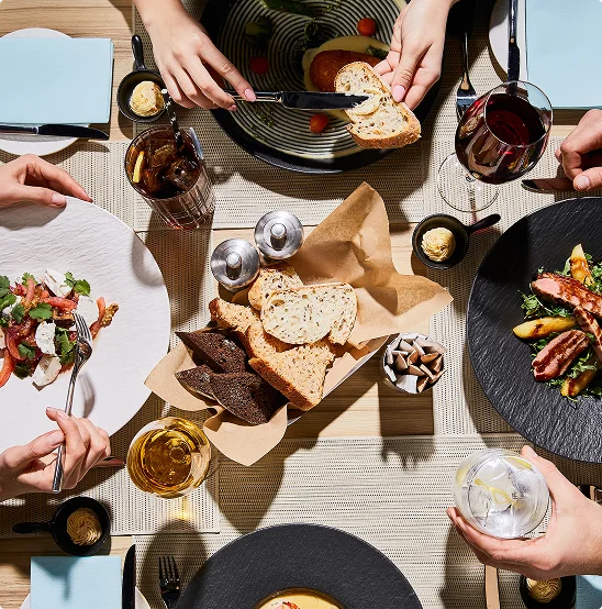 Een overzicht van een door zonlicht verlicht tafel waar meerdere handen reiken naar gesneden brood, salades en gerechten op donkere borden, omringd door glazen rode wijn, witte wijn en ijskoud water.
