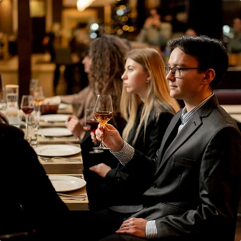 Een man in een grijs pak houdt een glas wijn vast aan een formeel gedekte restauranttafel, met andere gasten naast hem in een zacht verlichte eetkamer.