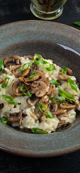 Een kom romige champignonrisotto, getopt met gebakken plakjes champignons en gehakte groene uien, wordt geserveerd in een rustieke keramische kom tegen een donkere achtergrond.