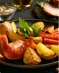 Een donkere koekenpan bevat geroosterde wortelgroenten — gouden aardappelen, oranje wortels en zoete aardappelpartjes — gegarneerd met een takje verse peterselie, met een hamgebraad zichtbaar op de achtergrond.