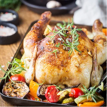 Een goudbruin hele geroosterde kip, gegarneerd met verse rozemarijn, rust in een donkere braadslee naast verkoolde tomaten, courgette en wortelgroenten op een houten oppervlak.