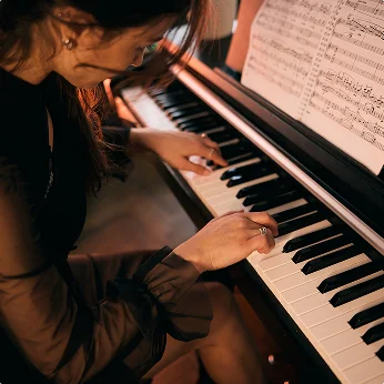 Een vrouw speelt op een vleugelpiano in warm, gedimd licht, haar handen op de toetsen met bladmuziek open op de standaard erboven.